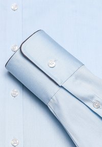 Camicia azzurro chiaro con una texture liscia, con bottoni bianchi e un bordo scuro a contrasto nell'area dei polsini e del colletto.