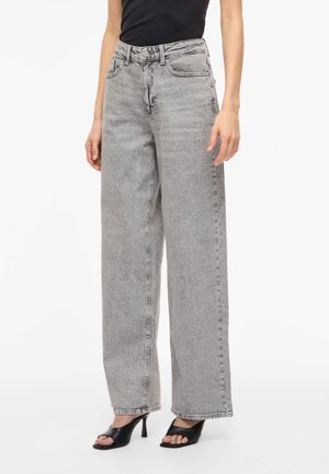 Femeie purtând blugi cu talie înaltă, de culoare gri deschis, cu picioare largi, asortați cu sandale negre cu toc înalt și vârf deschis și un top negru fără mâneci.