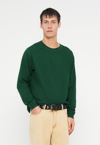 Groen lange mouwen crew neck shirt gemaakt van een gestructureerde stof, gedragen met lichtbeige denim broek en een zwarte riem met zilveren hardware.