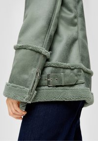 Close-up van een groene schapenvachten jas met een mouw, rits, gespdetails en een hand die rust bij donkerblauwe spijkerbroek.