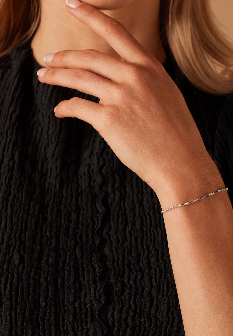 Silver bracelet on a wrist, featuring a delicate chain design, set against a textured black garment. Smooth skin with natural nails.