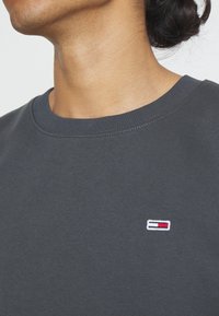 Gros plan d'une personne portant un t-shirt gris foncé à col rond avec un petit logo rectangulaire rouge, blanc et bleu sur la poitrine gauche.