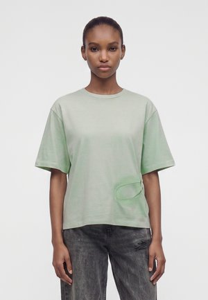 Giovane donna con capelli scuri raccolti che indossa una camicia larga verde chiaro a maniche corte e jeans grigio scuro su uno sfondo semplice.