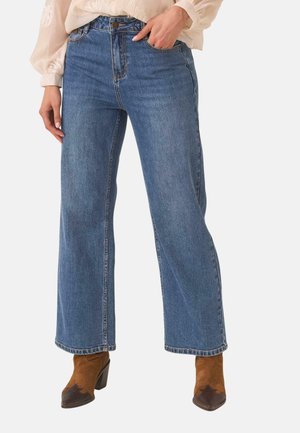 Mujer con vaqueros azules de talle alto y pierna ancha, con botón y cremallera, combinados con botas marrones de tacón y una blusa clara.