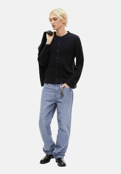 Jeune personne aux cheveux blonds portant un pull noir boutonné, un jean bleu clair avec des clés accrochées, des bottes noires, et tenant une veste sur l'épaule.