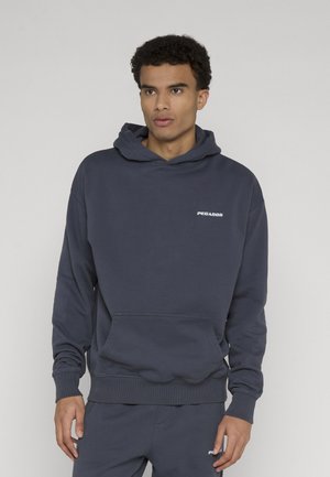 Hombre con sudadera con capucha azul oscuro y pantalones deportivos a juego, de pie frente a un fondo gris claro sencillo.