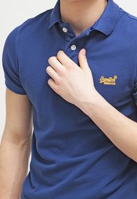 Mörkblå polotröja i bomull, med en tvåknappars placket och en orange Superdry-logotyp på vänster bröst. Korta ärmar.