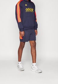 Námořnická mikina s pruhy na rukávech a kapse v červené a žluté barvě, s logem "adidas SPORTSWEAR". Kombinováno se sladěnými šortkami a šedými teniskami.