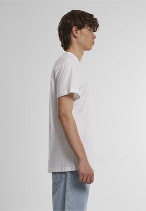 Młody mężczyzna z kręconymi włosami stoi bokiem, ubrany w jednolitą białą koszulkę i jasnoniebieskie dżinsy na neutralnym szarym tle.