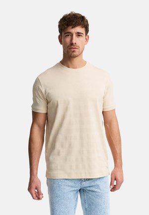Mand med kort krøllet hår iført beige tekstureret kortærmet skjorte og lyseblå jeans, stående mod en ensfarvet hvid baggrund.