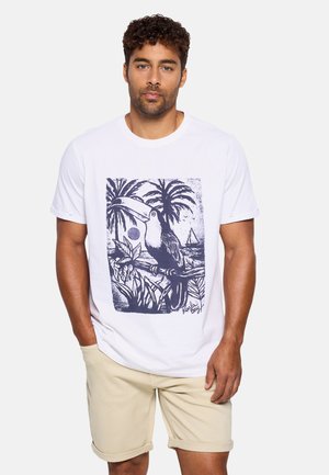 Hombre con camiseta blanca con un gráfico de tucán negro, palmeras y velero, combinado con pantalones cortos beige remangados.