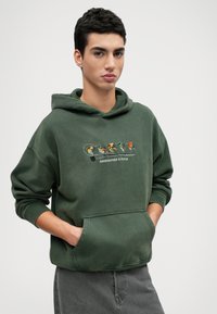 Zaļš džemperis no mīksta auduma, ar kabatu un izšūtām krāsainu putnu dizainiem. Brīva piegriezuma ar ribota materiāla aprocēm un apakšmalu.