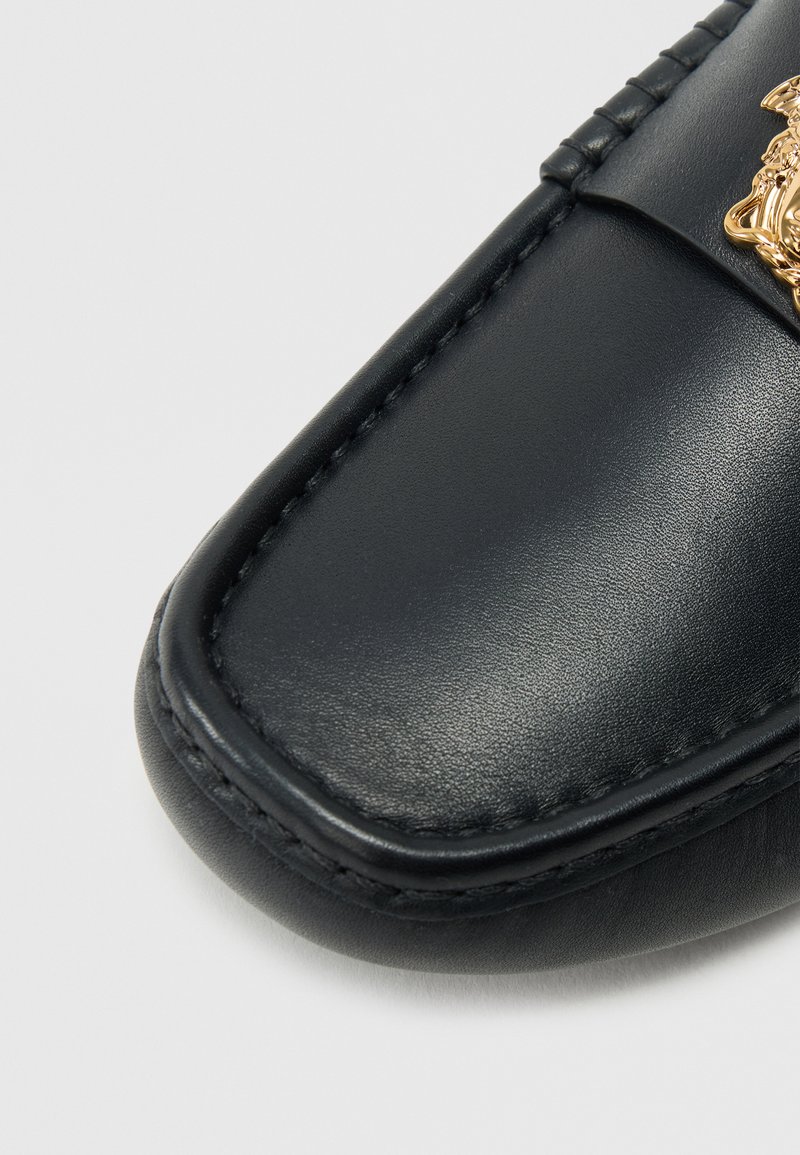 Schwarzer Lederloafer mit glatter Textur, rundem Zehenbereich und genähter Verzierung. Mit einem goldenen Emblem auf dem Oberteil.