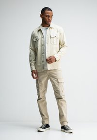 Beige overhemd met knopen en twee borstzakken, gecombineerd met beige cargobroek. Model draagt zwarte sneakers. Schone, gladde texturen.