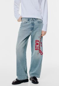 Ljusblå denimjeans med lös passform, med röd text "STWD" och stjärnor tryckta på sidan. Matchas med en vit långärmad tröja.