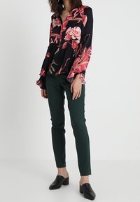 Blus med blommönster på svart bakgrund, utsvängda ärmar och knappar; kombinerad med gröna åtsittande byxor och svarta klackskor med blockklack.