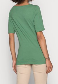 Zelené bavlněné tričko s krátkými rukávy, širokým výstřihem a rovným střihem, kombinované s béžovými kalhotami.