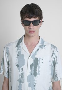 Camicia a maniche corte con colletto bianca e motivi astratti simili a vernice verde; modello che indossa occhiali da sole neri angolari.