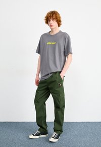 Grijze t-shirt met korte mouwen en groen logo, gecombineerd met ruime donkergroene cargobroek. Sneakers met witte zolen en zwarte veters.