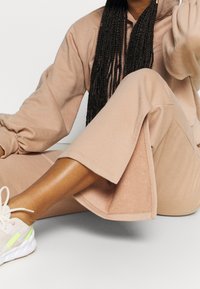 Persona seduta con le gambe incrociate, indossando pantaloni beige a gamba larga, felpa abbinata, sneakers bianche e lunghi capelli intrecciati con perline dorate.