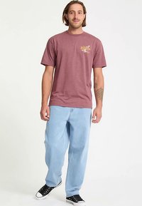 Bordeauxfarbenes Baumwoll-T-Shirt mit gelbem Grafiklogo, kombiniert mit hellblauen, locker sitzenden Jeans aus Denim und schwarzen Sneakers.