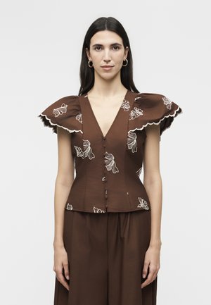 Γυναίκα που φοράει καφέ εφαρμοστή μπλούζα με λευκή κεντητή λουλουδένια διακόσμηση και μανίκια με κυματιστό τελείωμα, σετ με καφέ παντελόνι φαρδιάς γραμμής που ταιριάζει.