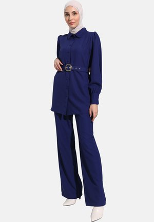 Blue top with button front and belted waist, paired with wide-leg pants. Made of textured fabric. Model wears a hijab and white heels.