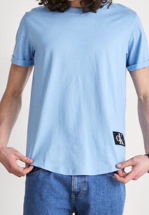 Osoba nosząca jasnoniebieską koszulkę z krótkim rękawem z podwiniętymi mankietami oraz małą czarną naszywką z logo Calvin Klein blisko dolnej krawędzi, w połączeniu z niebieskimi dżinsami.