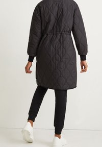 Giacca lunga nera trapuntata con vita aderente, polsini a costine, scollo rotondo e texture liscia. Indossata sopra pantaloni neri e sneakers bianche.