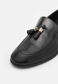 Mocassin en cuir noir avec des bords cousus et deux glands noirs maintenus par des anneaux métalliques dorés sur le dessus avant de la chaussure.