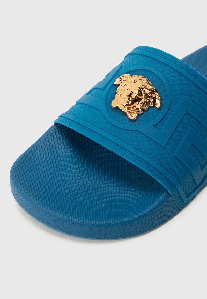 Blaue Gummisandale mit einer strukturierten Riemen, der ein goldfarbenes Medusa-Emblem und geometrische Muster zeigt. Glatte Innensohle für Komfort.