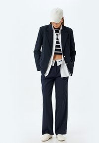 Marinblå blazer med silverknappar över en vit skjorta, randig kort topp och lösa marinblå byxor, kompletterat med en krämfärgad keps och vita sneakers.