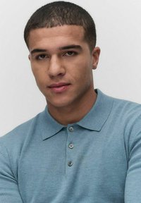 Ljust blått stickat polo shirt med krage, med tre knappar och subtil textur, bärs av en person med kort hår.