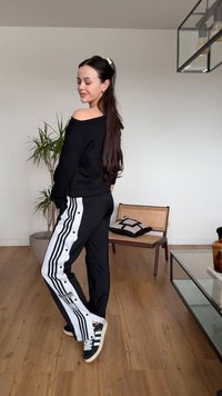 Vrouw met lang donker haar in een zwarte off-shoulder top en Adidas trainingsbroek die binnen poseert bij een plant en een houten stoel met kussens.