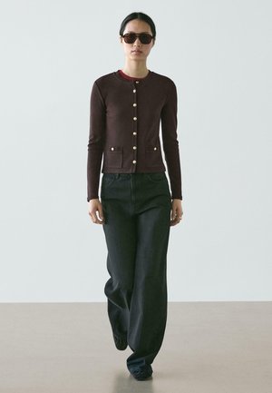 Žena kráčející s tmavými slunečními brýlemi, vínově červenou bundou zapnutou na zlaté knoflíky, černými kalhotami s širokými nohavicemi a černými botami na jednobarevném pozadí.