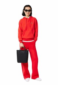Sweat à capuche rouge avec poche avant, associé à un pantalon de survêtement rouge assorti. Sac à main noir avec deux poignées et détails de logo. Baskets blanches.