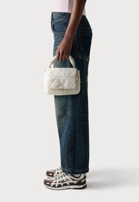 Hvid quiltet håndtaske lavet af blødt materiale med en trinaktig accent og guldfarvet hardware, holdt i en persons hånd, parret med denim jeans.