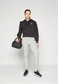 Black zip-up sweatshirt, gray sweatpants, black sneakers, and a black bag. Smooth fabric with a fitted shape. Logo detail on the hoodie.