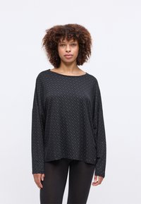 Mulher com cabelo encaracolado, vestindo uma blusa preta de mangas longas e larga com padrão e leggings pretas, de pé contra um fundo branco liso.