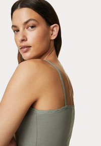 Jasnozielona koszulka na ramiączkach z koronkowym wykończeniem i cienkimi paskami, uszyta z miękkiego materiału. Posiada dopasowany krój, który podkreśla ramiona.