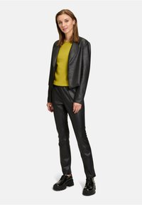 Zwarte neplederen cropped blazer en strakke broek, gecombineerd met een levendige felgroene trui en stevige zwarte schoenen. Glad oppervlak, op maat ontworpen.