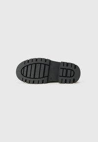 Semelle de chaussure en caoutchouc noir avec des rainures profondes et des motifs de bande de roulement horizontaux et verticaux, présentée sur un fond gris clair uni.