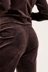 Hnedé velúrové teplákové nohavice majú priliehajúci pás a na zadnej strane sa nachádza logo "Juicy" posiate kryštálmi, s hladkou a jemnou textúrou.