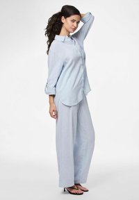 Svijetloplava, široka pamučna košulja s dugmadi i rukavima na preklop, u kombinaciji s odgovarajućim širokim hlačama i tankim sandalama.