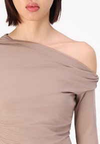 Cămașă off-the-shoulder dintr-o țesătură moale, de culoare bej deschis, cu drapaje delicate, având o croială ajustată și o textură netedă.