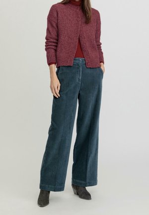 Femme portant un cardigan bordeaux boutonné, un pull col roulé couleur rouille, un pantalon large en velours côtelé bleu et des bottines foncées, debout les mains dans les poches.