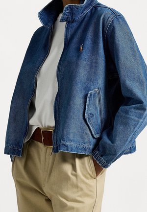 Man draagt een blauwe spijkerjack met ritssluiting over een lichtgrijs shirt, beige broek met een bruine riem, en heeft zijn handen in zijn zakken.