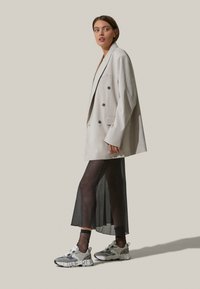 Blazer beige ajustado con botones de doble botonadura, combinado con una falda negra semitransparente y zapatillas deportivas grises con detalles de malla y texturizados.