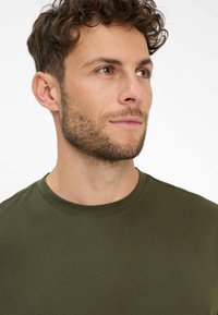 Olivgrünes Rundhalsausschnitt-T-Shirt aus glattem Stoff; mit kurzen Ärmeln und einem taillierten Design mit dezenten Stichdetails.