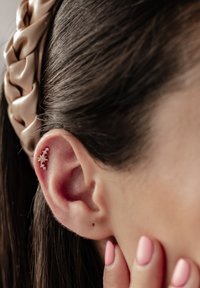 Boucle d'oreille en forme d'étoile dorée avec des accents transparents, présentée dans l'oreille. Les cheveux sont noirs et lisses, avec un bandeau tressé beige clair. Les ongles sont rose clair.